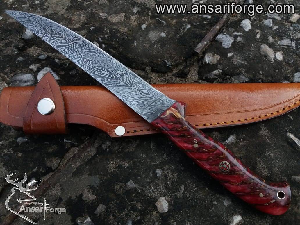 A fish fillet knife with a Damascus steel blade, a red pine cone handle, and a leather sheath next to a pile of fresh fish fillets.