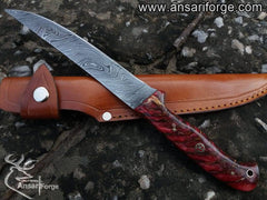 A fish fillet knife with a Damascus steel blade, a red pine cone handle, and a leather sheath next to a pile of fresh fish fillets.